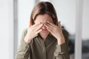 A woman wearing a brown shirt with her hands on her face in front of a window
