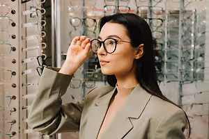 A woman is standing in front of a glass display of eyeglasses and is looking through a pair of eyeglasses with her right hand.