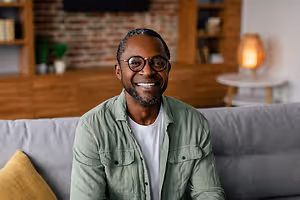 Black man wearing glasses and green shirt sitting on a couch with a smile on his face.