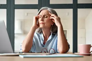 An older woman sitting at a desk with her hands on her head, looking tired and holding a cup of coffee.