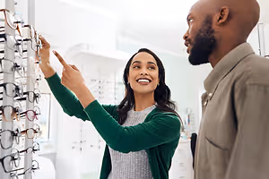 A woman in a green cardigan points to a pair of glasses in a shop while smiling, with a man looking at her. Behind them, a wall with a picture frame and a white shelf with glasses is visible.