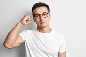 A man wearing glasses and a white t-shirt is posing for a photo, with a hand on his forehead.