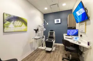 A medical office with a blue wall, a desk with two monitors, a mounted monitor, a framed certificate, and a black chair near a medical chair with a white tray, all illuminated by ceiling lights.