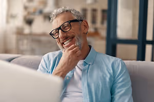 An older man wearing glasses is smiling while sitting on a couch and looking at a laptop.