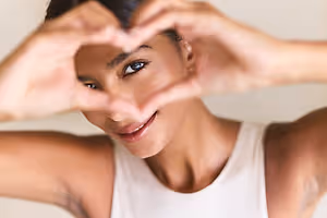 Woman wearing white tank top and smiling while making a heart shape with her hands