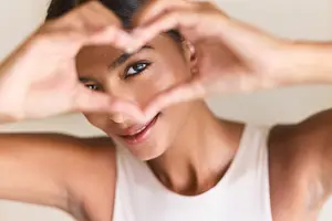 Woman wearing white tank top and smiling while making a heart shape with her hands