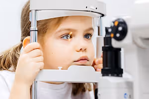 A young girl with brown hair is looking through an eye examination machine, likely in a clinic.
