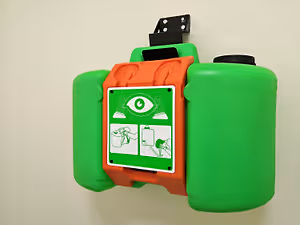 An eye wash station with a green and red body, mounted on a wall, displaying an icon with a green circle and an eye symbol, and two illustrations showing a person washing their eyes and another person using a bottle