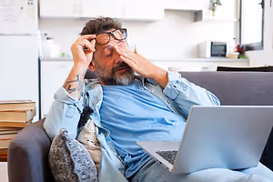 A man is lying on a couch with his hand covering his eyes and a laptop on his lap.