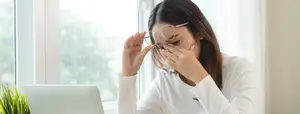 Woman sitting by a window with her hands covering her face, possibly sneezing.