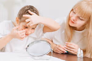 Two women sitting at a table with one holding a mirror while the other puts eye drops in her eyes.