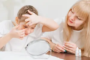 Two women sitting at a table with one holding a mirror while the other puts eye drops in her eyes.