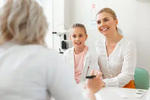 An adult woman and a young girl are sitting in front of a woman in a white coat, who is smiling and looking at them. The adult woman is holding a pen and paper, while the young girl is holding a tablet. Behind them is a wall with an eye chart, and a chair is on the right side.