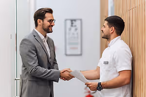 Two men in suits, one holding a paper, shaking hands in a modern office setting.
