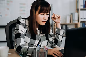 An East Asian woman working on a laptop with a cup on the desk and a plant in a cup holder.