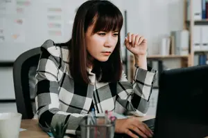 An East Asian woman working on a laptop with a cup on the desk and a plant in a cup holder.