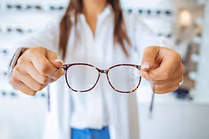 A woman is holding a pair of eyeglasses with brown frames.