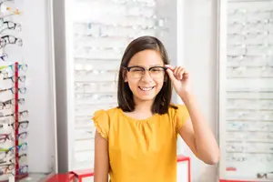 A young girl wearing glasses stands in front of a display of eyeglasses in a store.