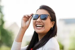 A woman adjusts her sunglasses and smiles at the camera, with a blurred background of a building and trees.