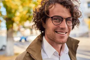 A smiling man with curly hair wearing glasses and a brown jacket is standing on the sidewalk in front of a tree and a parked car.