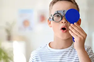 A boy is wearing a pair of blue-framed eyeglasses and holding a blue lens in his hand, possibly trying to see through it while looking upward.