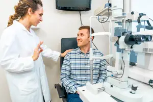 A doctor is talking to a man in a clinic as he sits in front of an eye examination machine.