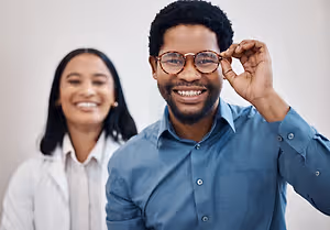 A man and a woman are standing together, the man is wearing glasses and smiling, and the woman is standing behind him with a smile