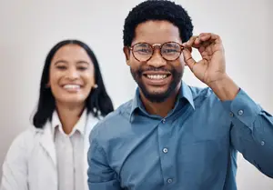 A man and a woman are standing together, the man is wearing glasses and smiling, and the woman is standing behind him with a smile
