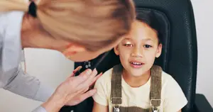 A woman is examining a young girl's eyes with a tool, with a black chair in the background.