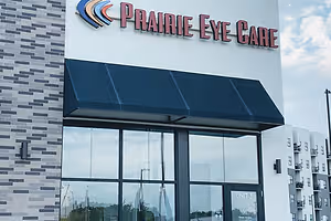 A building with a black awning with the words 'Prairie Eye Care' on it. The building has a brick wall on the left side and glass windows on the right side. The glass windows reflect the surroundings. There is a sign with the text 'UNIT 3' on the door. The sky is cloudy.