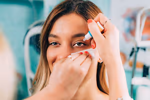 A woman is applying a white cream to her eyebrow area with a stick, while another person is holding her face with their hand. The woman is wearing a ring, bracelet, and necklace. Behind her is a blurry background with a white object, possibly a chair, and a green wall.