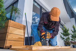 A person drilling on a wooden plank on a wooden floor near a building