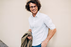 man with glasses and a white shirt holding a brown jacket in his left hand and smiling