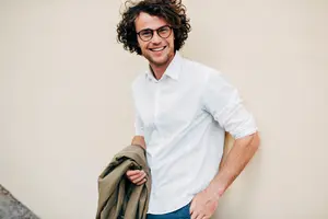 man with glasses and a white shirt holding a brown jacket in his left hand and smiling