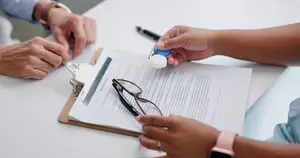 A man and a woman are sitting at a table reviewing a document, the woman is holding a pair of glasses and a contact lens bottle.