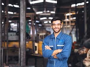 A man in a blue jumpsuit stands with his arms crossed in a factory setting