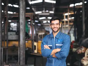 A man in a blue jumpsuit stands with his arms crossed in a factory setting
