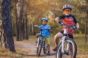 Two kids riding bicycles on a forest trail with trees in the background