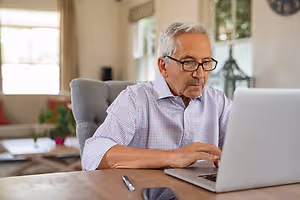 An older man wearing glasses is sitting at a desk and typing on a laptop