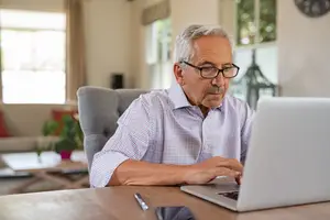 An older man wearing glasses is sitting at a desk and typing on a laptop