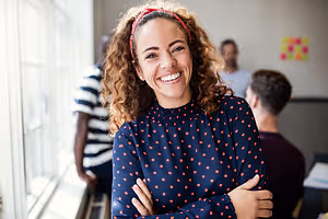 A woman with curly hair wearing a red headband is smiling and posing for a photo while standing in front of a window. Behind her, a man is sitting and looking at something on the right side. On the left side, a person is standing and looking at something. On the wall, there is a yellow and orange sticker.