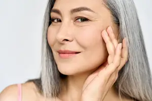 A woman with gray hair is smiling and touching her face with her right hand, wearing a pink strap on her shoulder. She is probably posing for a photograph in a studio.