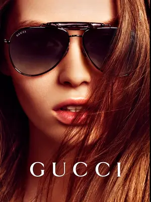 Close-up of a woman wearing Gucci sunglasses with brown lenses and silver frames, the brand name is displayed prominently in white letters at the bottom of the image