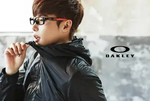 A young person wearing a black jacket and Oakley glasses stands against a gray wall