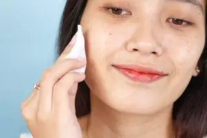 A woman with a ring on her left hand, holding a white cloth, and smiling while applying makeup on her face.