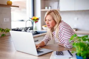 An older woman is working on her laptop while leaning against a counter in her kitchen.