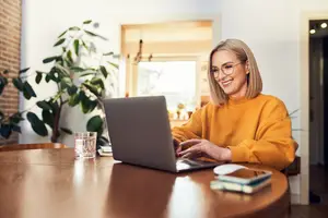 A woman wearing glasses and a yellow sweater sitting at a table with a laptop smiling