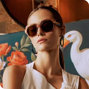 A woman in a white dress wearing brown sunglasses and large hoop earrings poses in front of a painting of a swan and flowers.