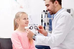 A smiling male optometrist is examining a girl patient's eyes with an ophthalmoscope in a clinic