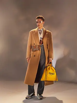 A man wearing a beige coat and eyeglasses with a Fendi bag and a belt around his waist standing in a studio